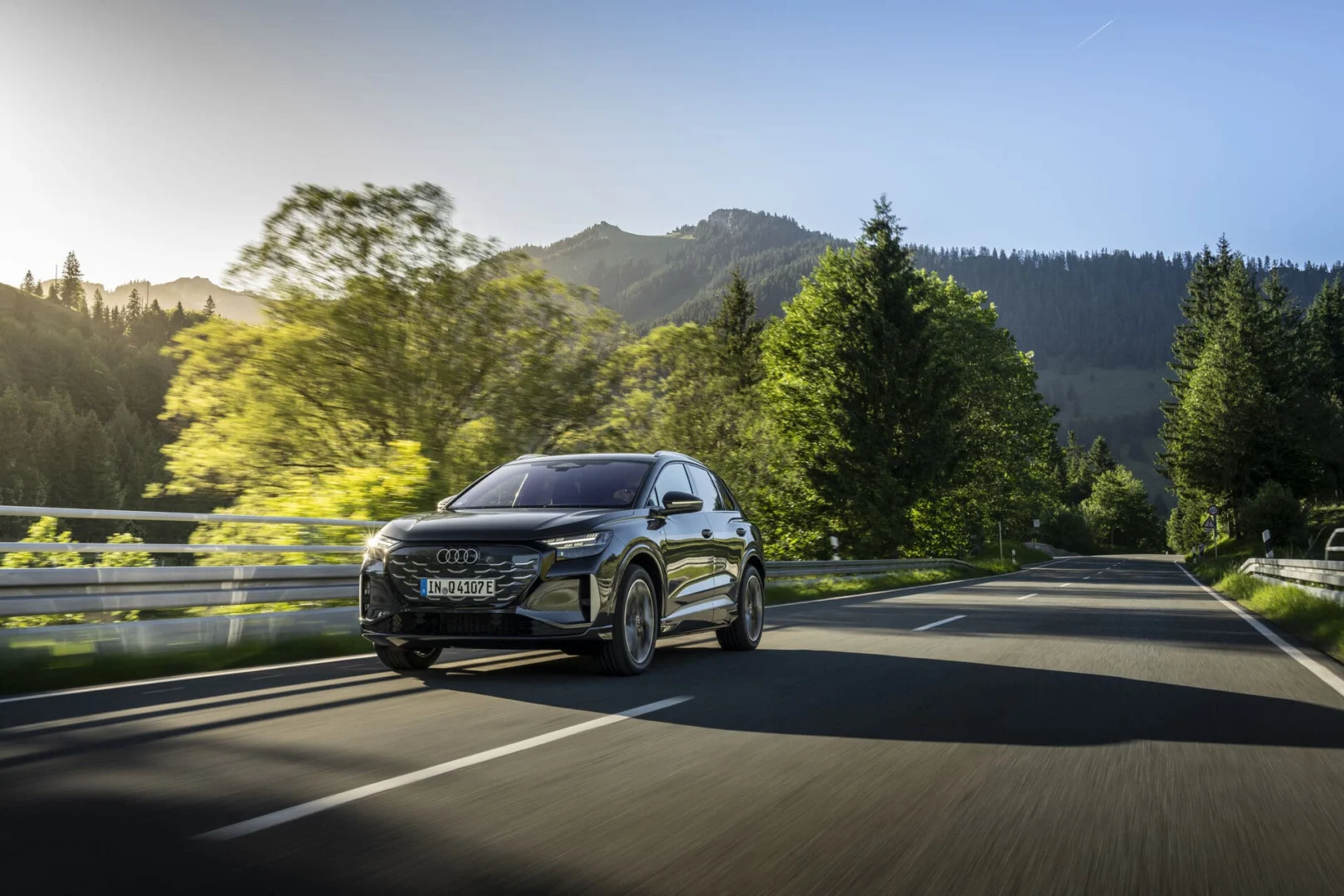 The right car for any situation: the Audi Q4 e-tron takes to the streets - Audi Q4 SUV e-tron in Tamboragrey, outdoor driving shot, exterior, three-quarter front view on a country road surrounded by forest and mountain scenery.